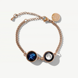 a rose gold bracelet featuring two charms, one with a moon phase image and the other with a constellation image, set against a white background.